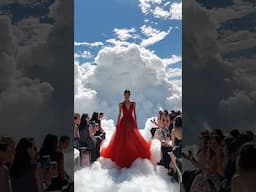 A model in a flowing red gown glides across a cloud filled runway